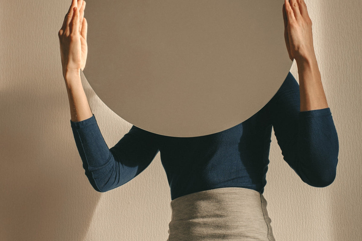 Woman holding a mirror up facing away from her on neutral background.