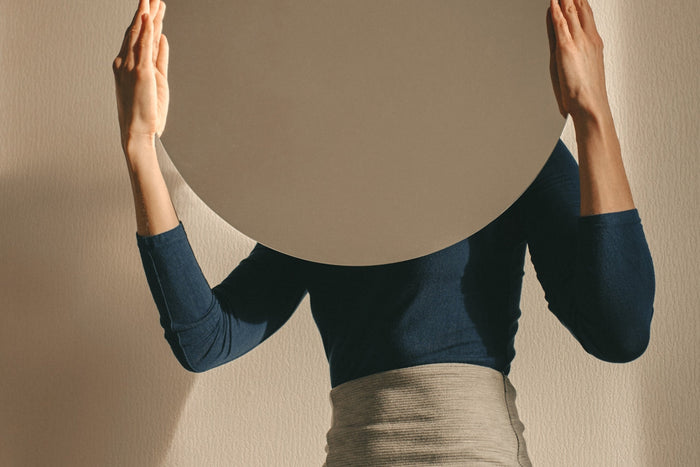 Woman holding a mirror up facing away from her on neutral background.