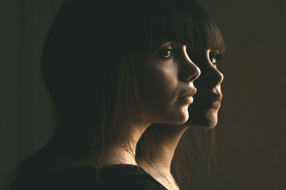 Editorial image of two identical women with thoughtful expressions against a dark background with soft lighting.