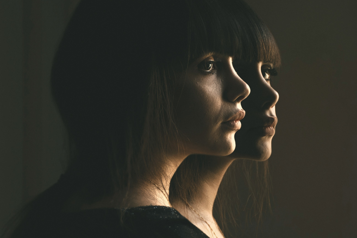 Editorial image of two identical women with thoughtful expressions against a dark background with soft lighting.