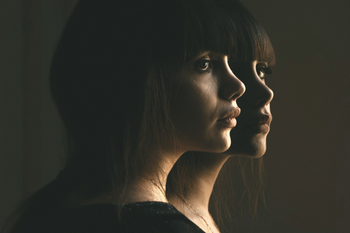 Editorial image of two identical women with thoughtful expressions against a dark background with soft lighting.