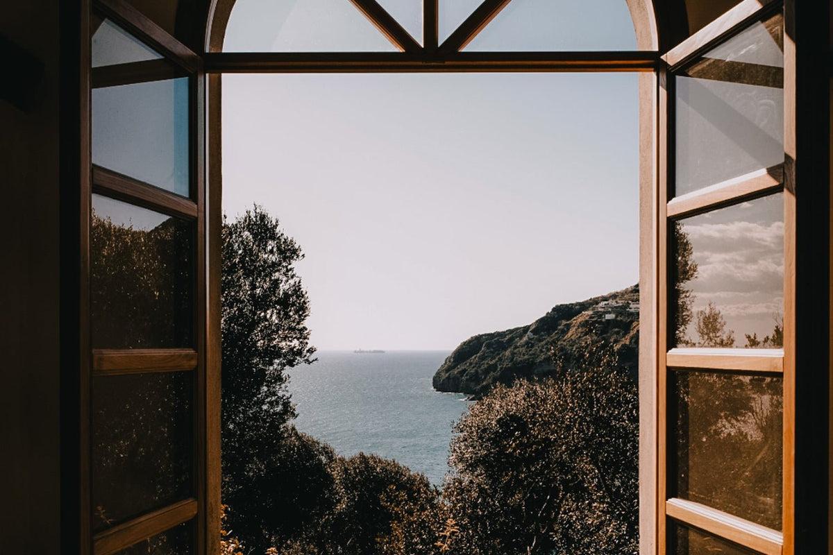 An open window overlooking a blue ocean with greenery.