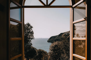 An open window overlooking a blue ocean with greenery.