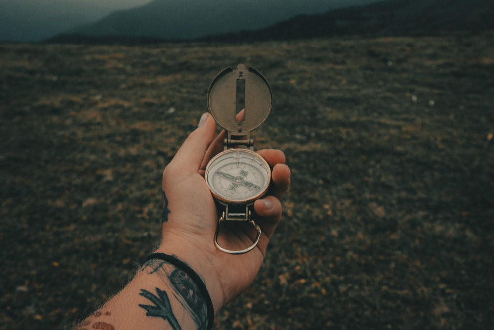 A hand holding a compass with no clear direction against mountainous terrain.
