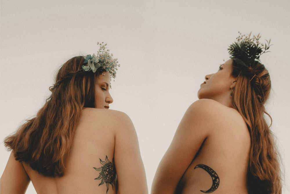 Two twin women with floral crowns and matching sun and moon tattoos outdoors under gray skies.