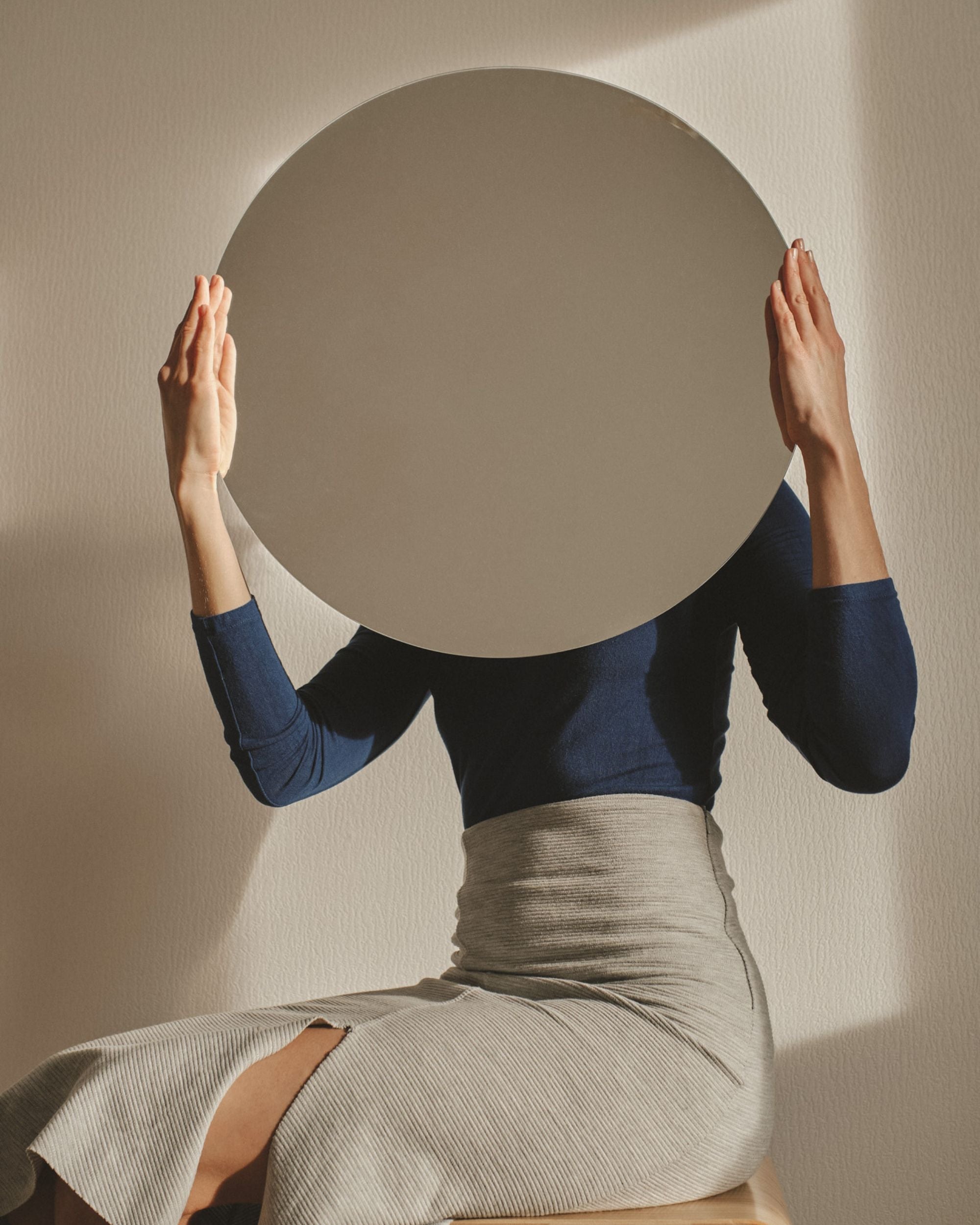 Woman holding a mirror up facing away from her on neutral background.