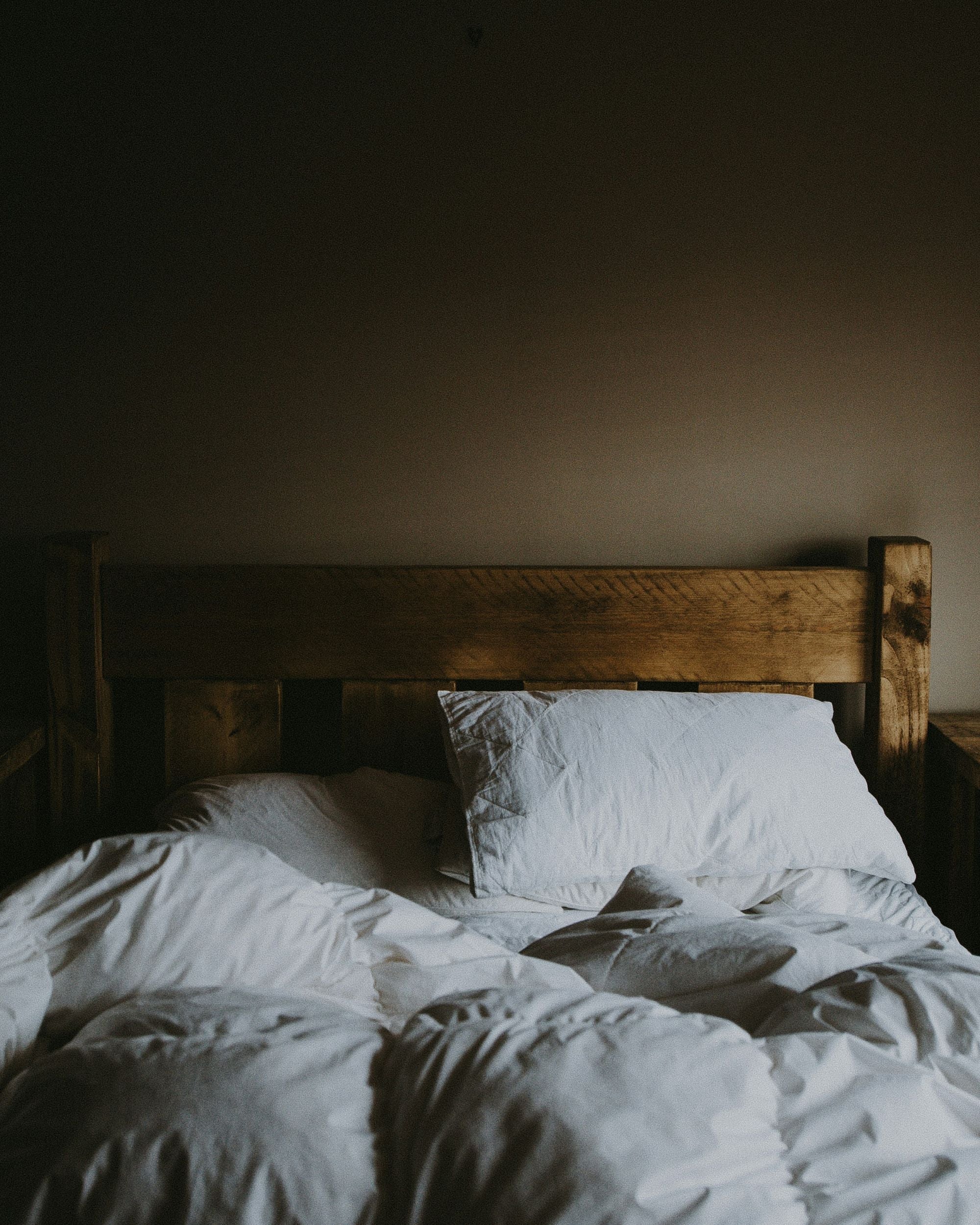 Unmade bed with a wooden frame and white sheets against a neutral beige wall.