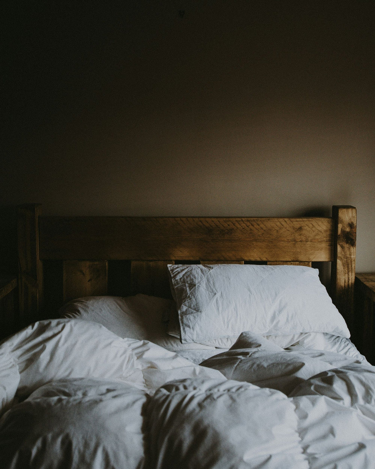 Unmade bed with a wooden frame and white sheets against a neutral beige wall.