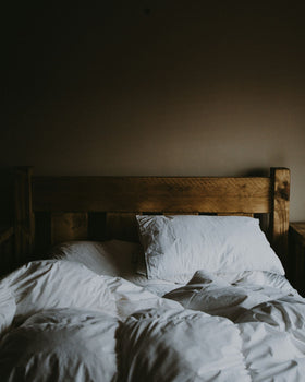 Unmade bed with a wooden frame and white sheets against a neutral beige wall.