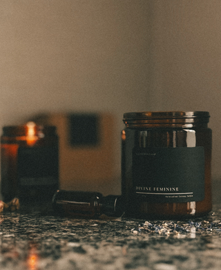Amber glass candle labeled "Divine Feminine" by Alchemist + Co, styled on stone with essential oil bottle and dried botanicals, evoking ritual and warmth.