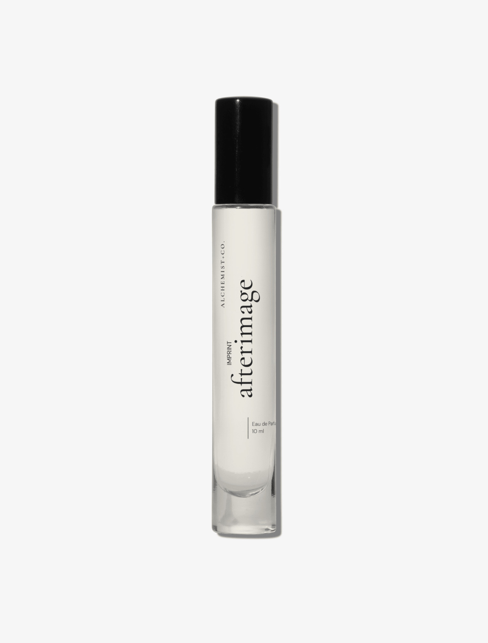 A clear glass perfume bottle with a black cap, featuring a white label with the brand name 'Afterimage' by Alchemist + Co.  set against a solid light gray background.