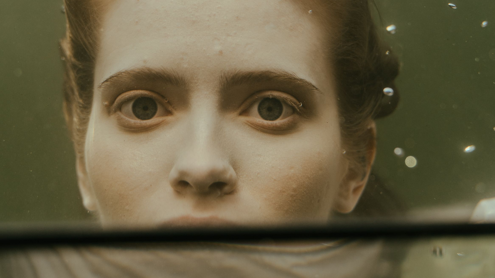 A close-up portrait of a woman underwater, with her face partially obscured by the waterline and a dark horizontal band across the lower half. Her hair floats upward through the water, and the scene has a muted olive-green cast with visible air bubbles.