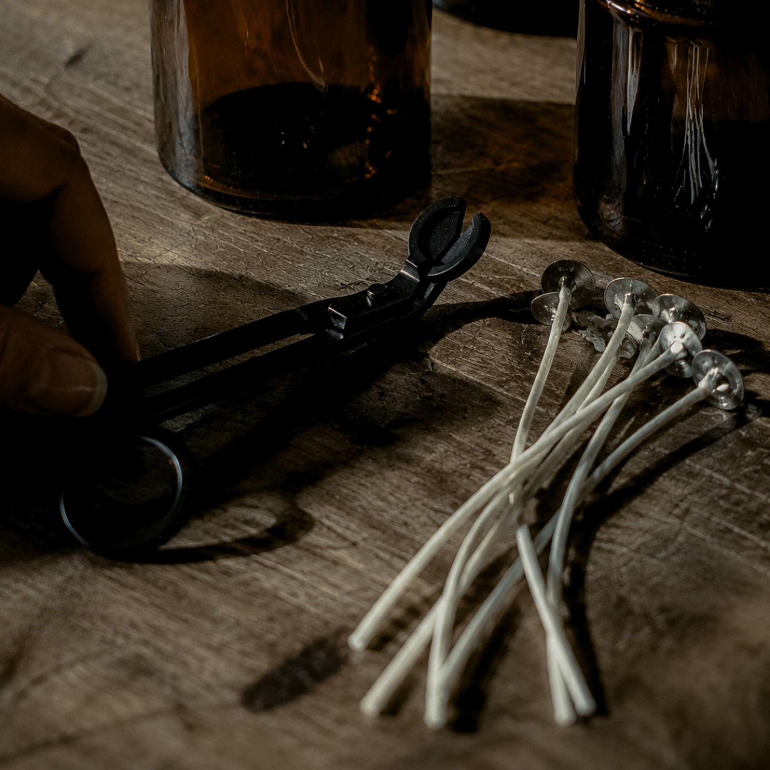 Black wick trimmer and a bundle of cotton wicks on a wooden surface with empty amber candle jars in the background