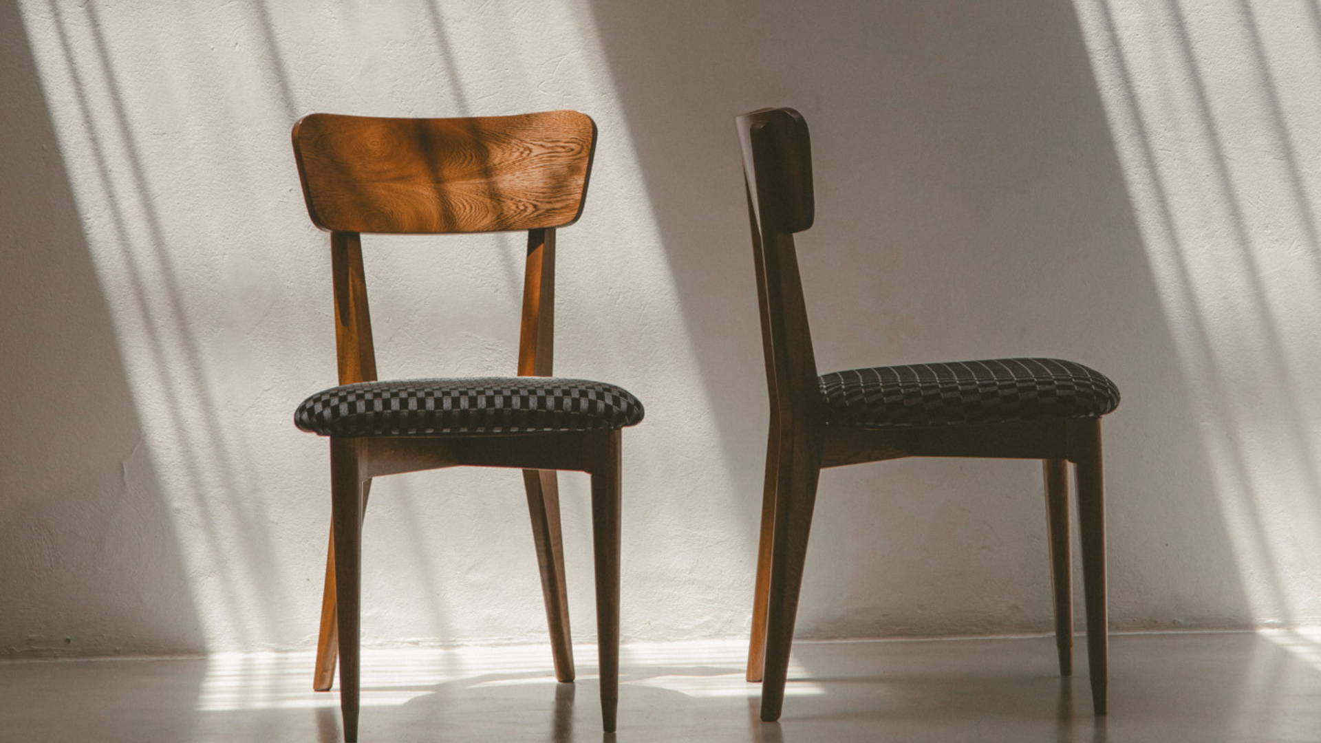 Two modern wooden dining chairs with patterned upholstered seats, standing against a white wall with dramatic diagonal sunlight shadows.
