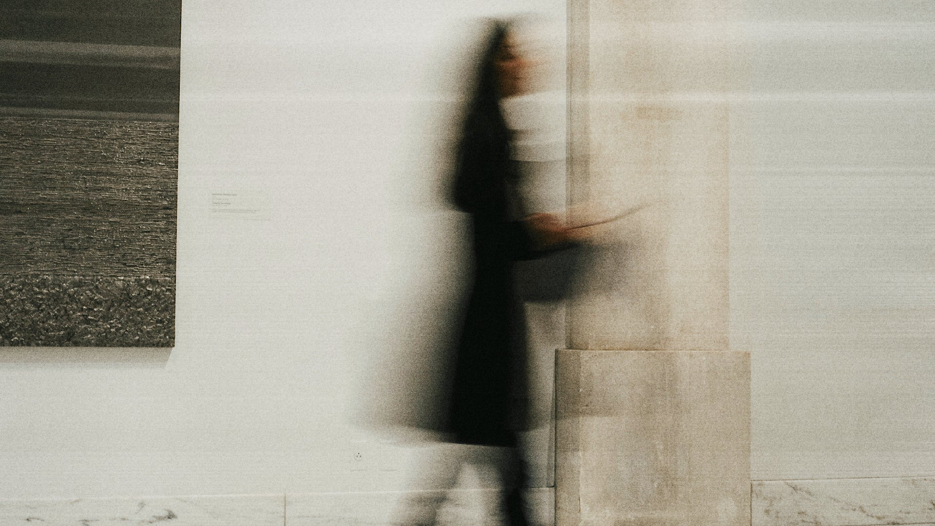 A blurred figure in a dark coat walks past a light-colored wall in an indoor setting, with a framed dark artwork visible on the left.