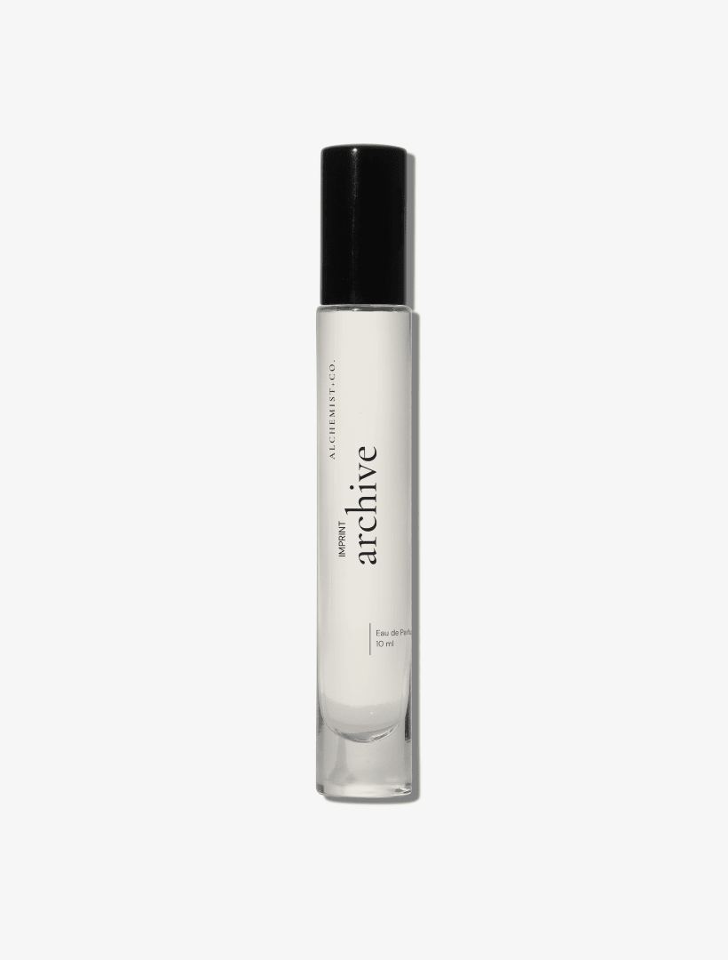 A clear glass perfume bottle with a black cap, featuring a white label with the brand name 'Archive' by Alchemist + Co.  set against a solid light gray background.