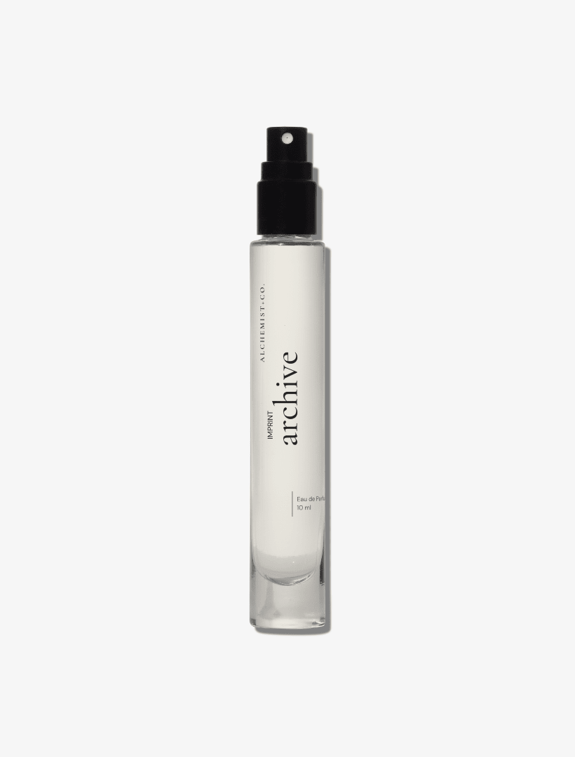 A clear glass perfume bottle featuring a white label with the brand name 'Archive' by Alchemist + Co.  set against a solid light gray background.
