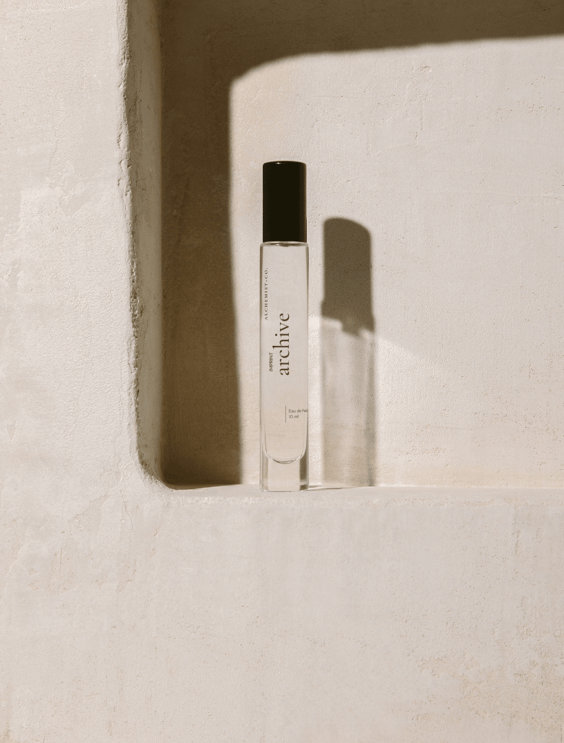 A clear glass perfume bottle with a black cap, featuring a white label with the brand name 'Archive' by Alchemist + Co.  set against a stone background.