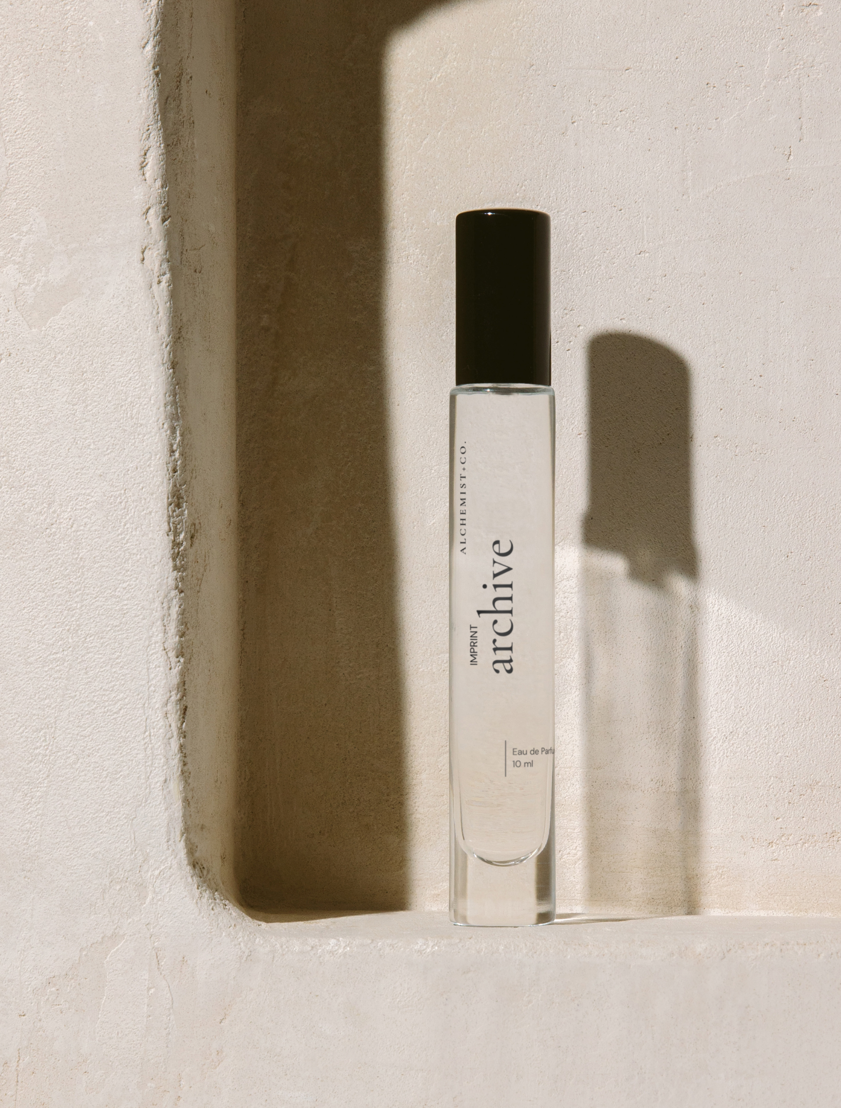 A clear glass perfume bottle with a black cap stands on a textured beige surface in a recessed niche, casting a sharp shadow. The bottle is labeled 'Archive' by Alchemist + Co. 