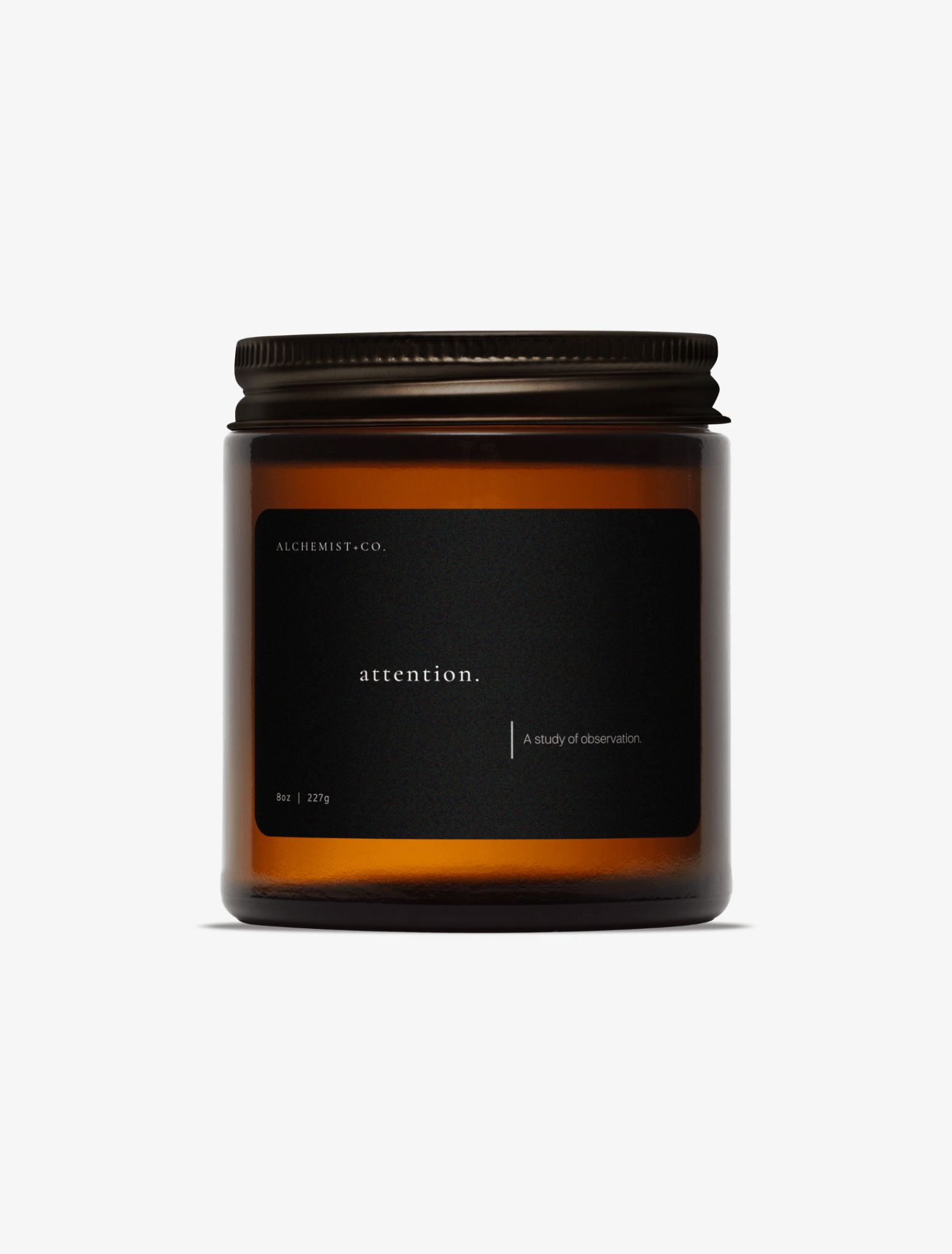 An amber glass jar candle with a black label and a dark metal lid, centered on a light gray background. The label reads 'Attention'.