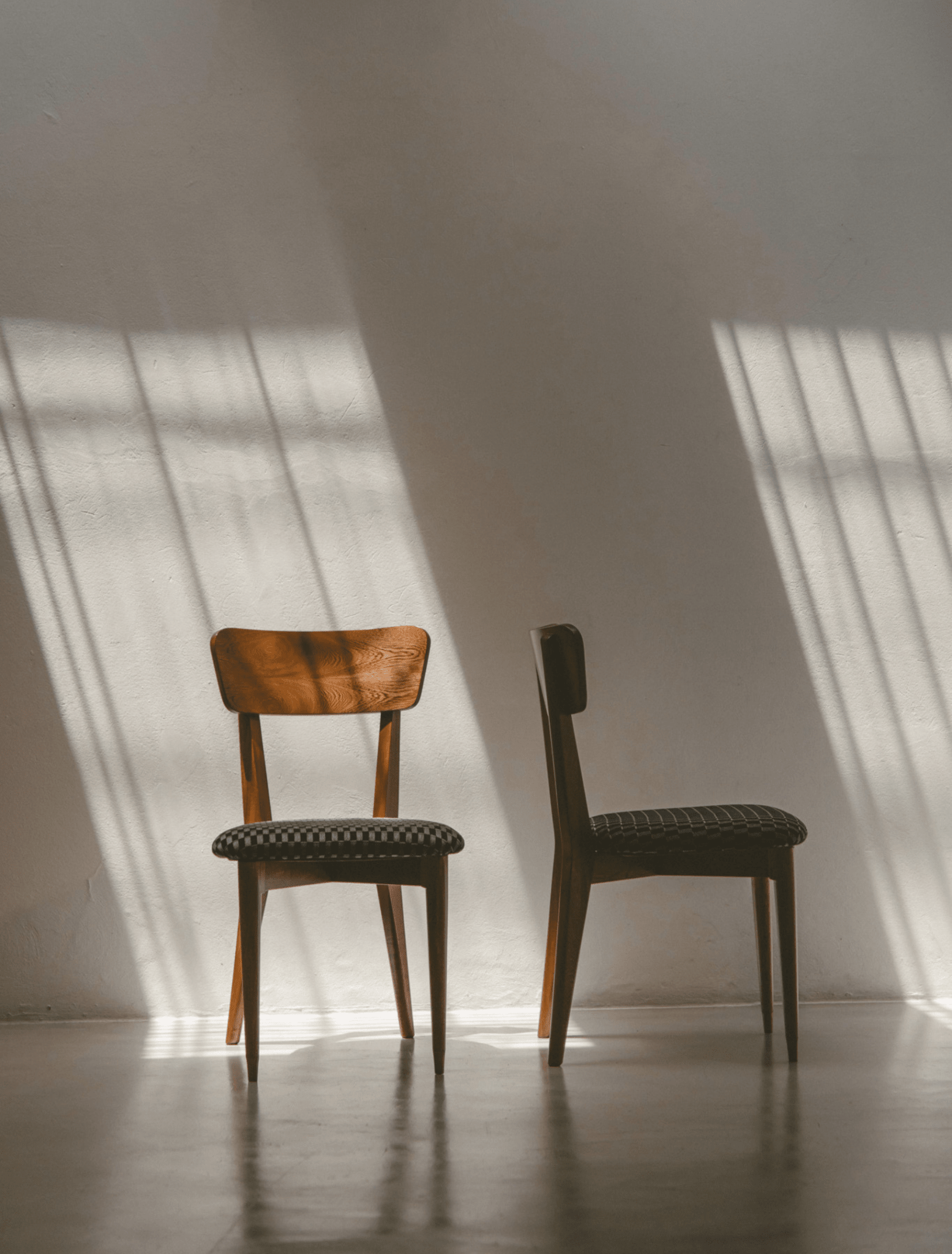 Two wooden dining chairs with dark upholstered seats sit side by side in a minimalist room with dramatic window light casting shadows on a white wall and concrete floor.