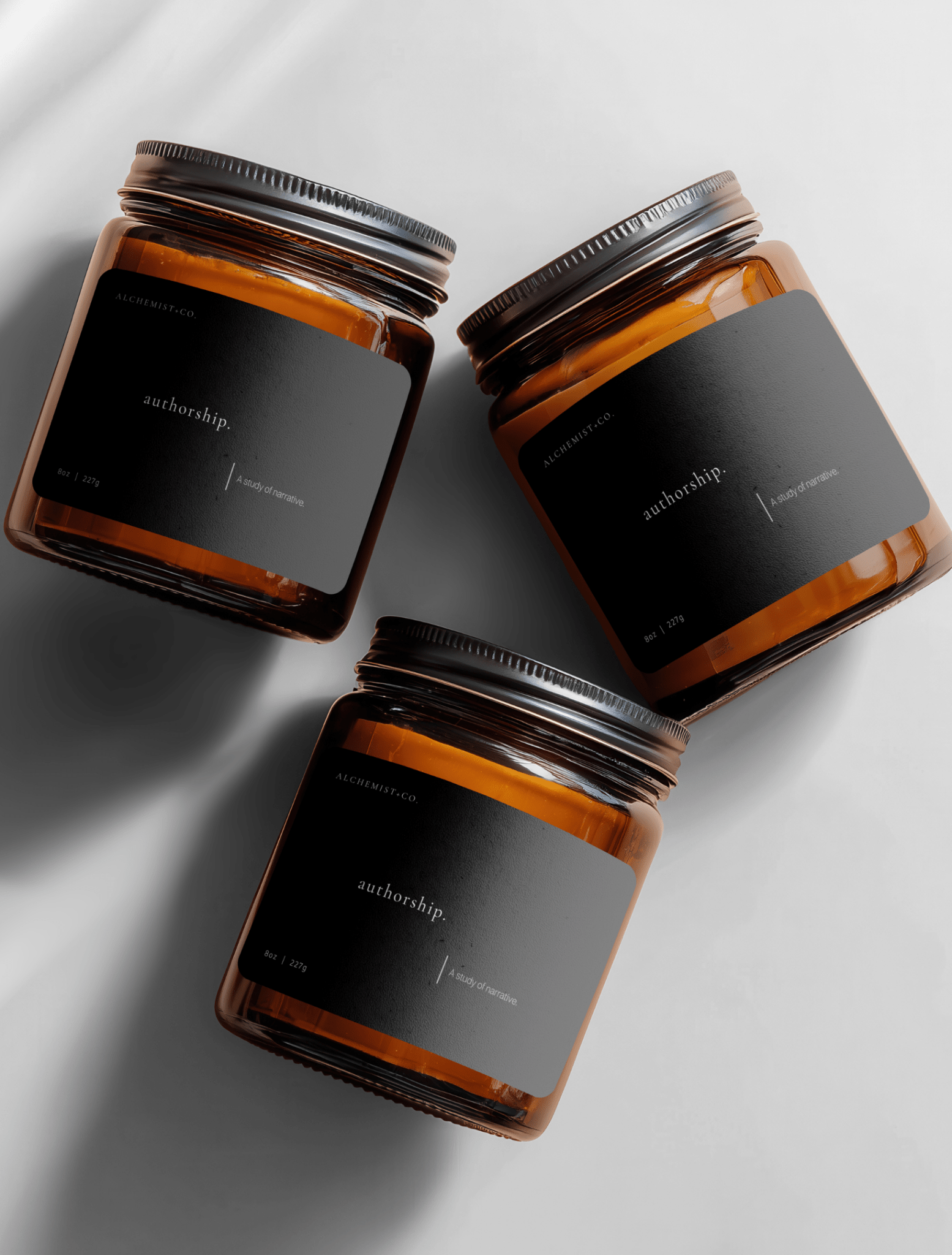 Three amber glass jar candles with black labels and silver lids, arranged diagonally on a light gray background with soft shadows labeled 'Authorship'.