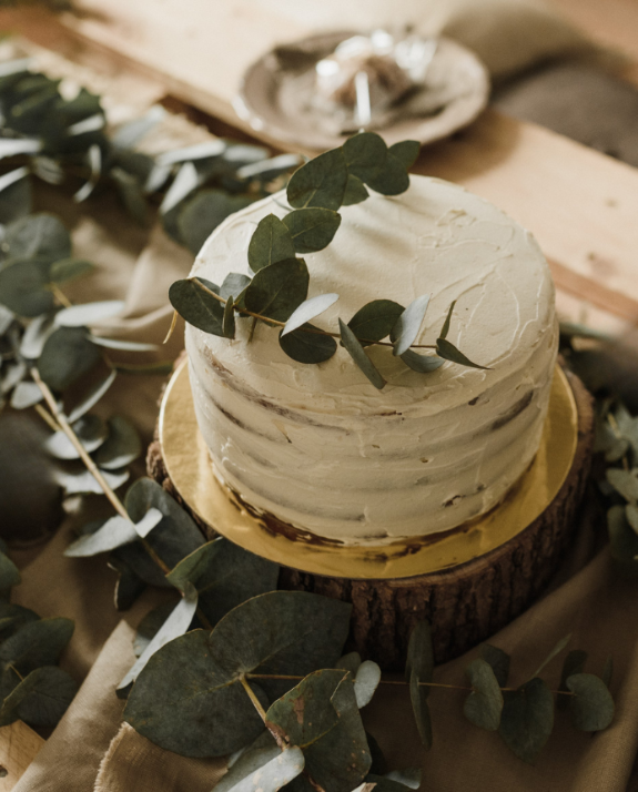 White frosted cake with greenery on a wooden stand, surrounded by eucalyptus leaves.