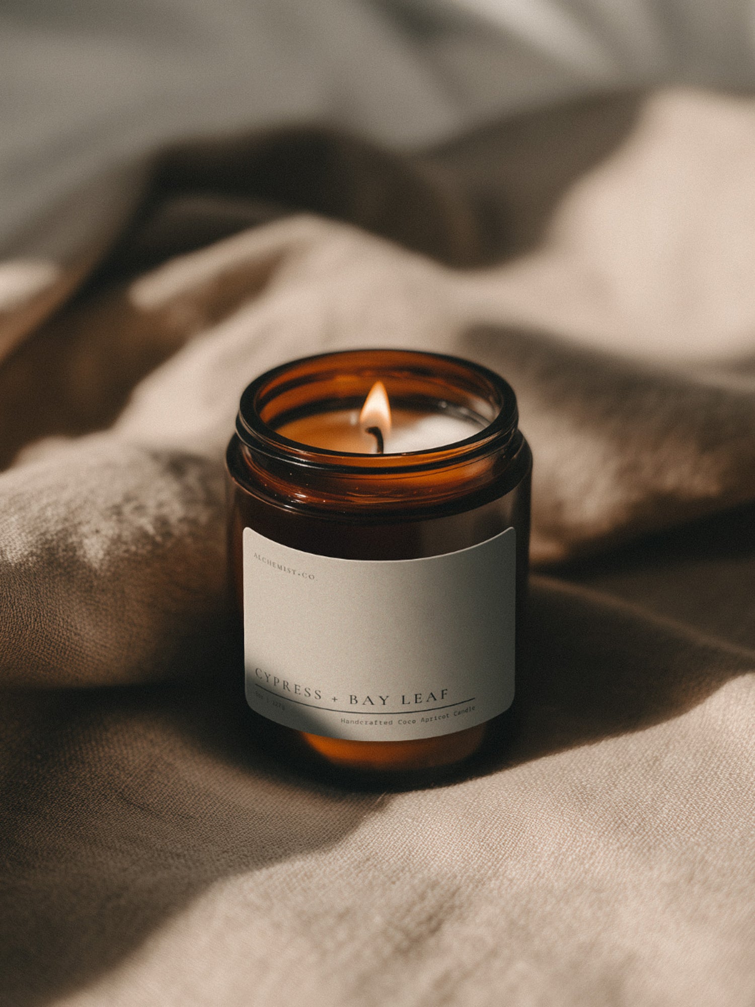 Cypress + Bay Leaf Bare candle burning beside soft linen in warm atmospheric light.