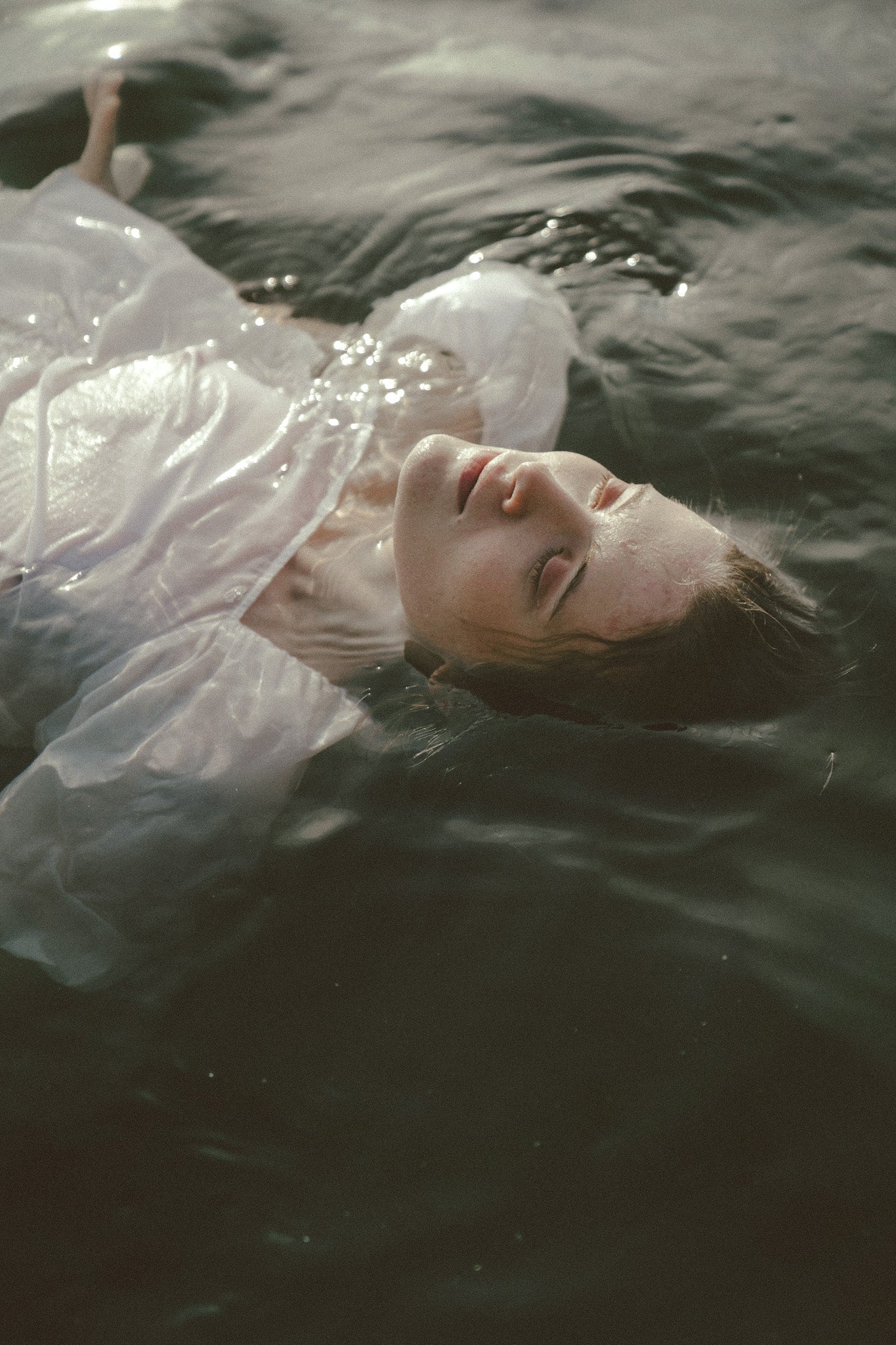 A person floating on their back in dark water, wearing a sheer white translucent top or dress. The face is partially obscured by water and ripples, with a moody, low-light aesthetic.