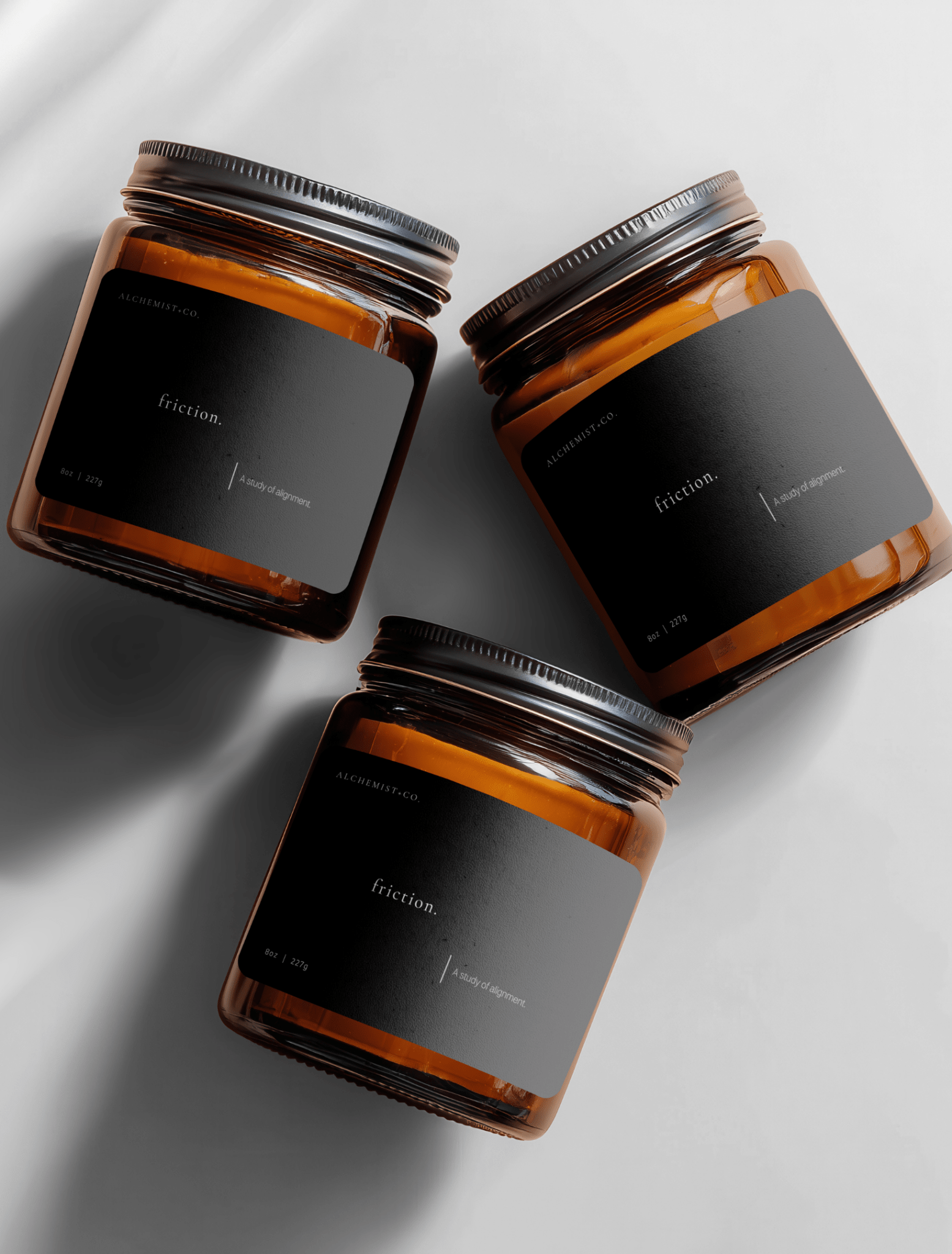 Three amber glass jar candles with black labels and silver lids, arranged diagonally on a light gray background with soft shadows labeled 'Friction'.