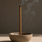 Hand Rolled Palo Santo incense stick burning in a ceramic bowl with smoke against a dark background.