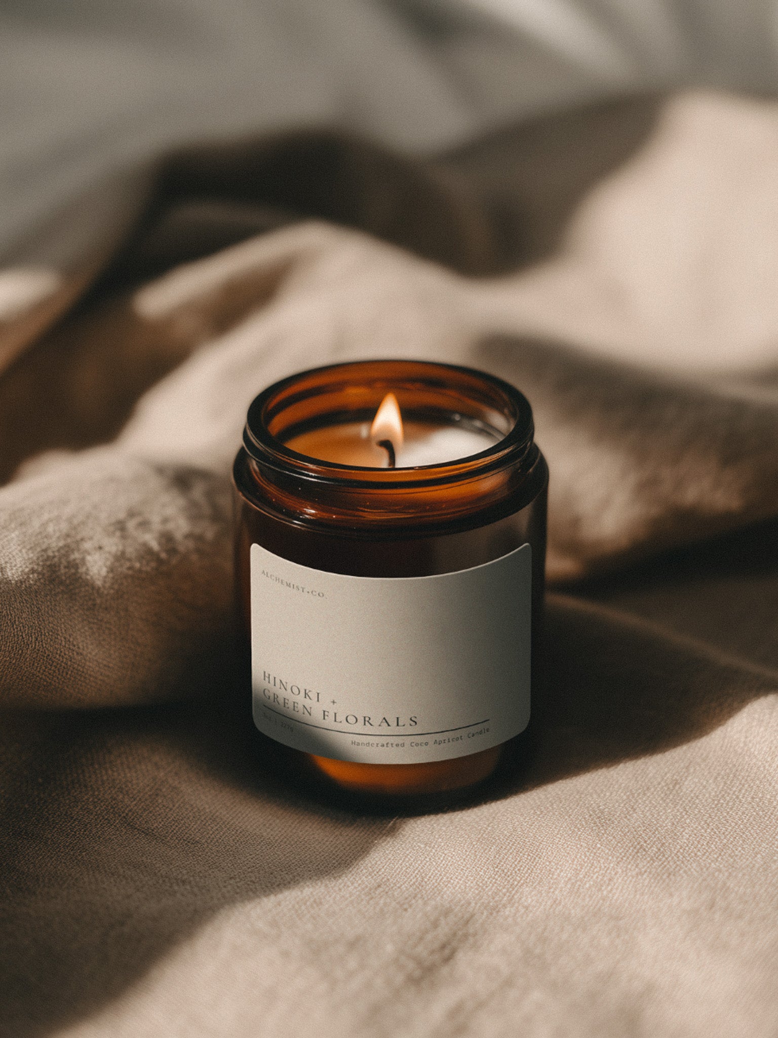 Hinoki + Green Florals Bare candle burning beside soft linen in warm atmospheric light.