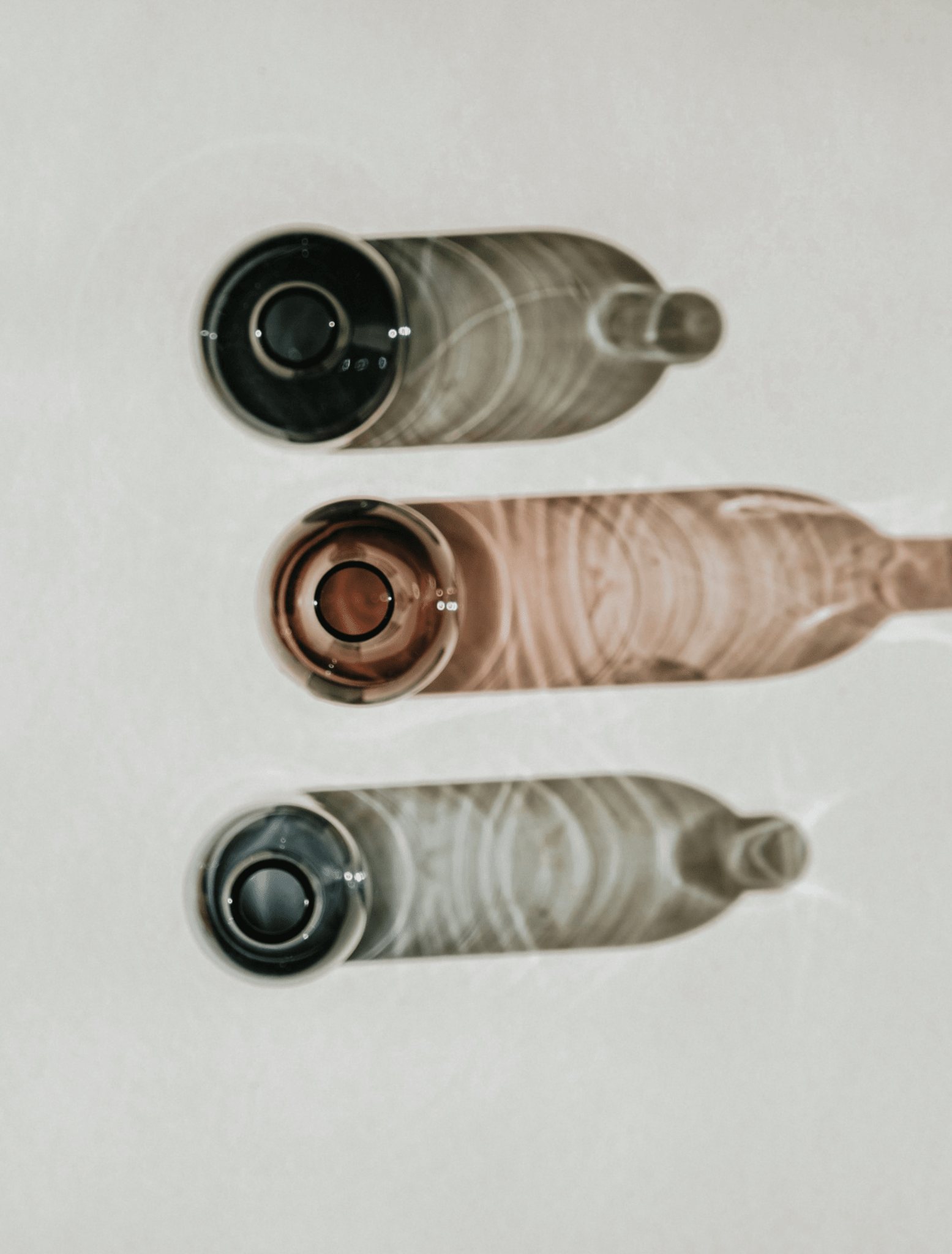 Three glass bottles vertically on a light off-white background. The top bottle is dark gray/black, the middle bottle is a dusty rose pink, and the bottom bottle is a smoky translucent gray.