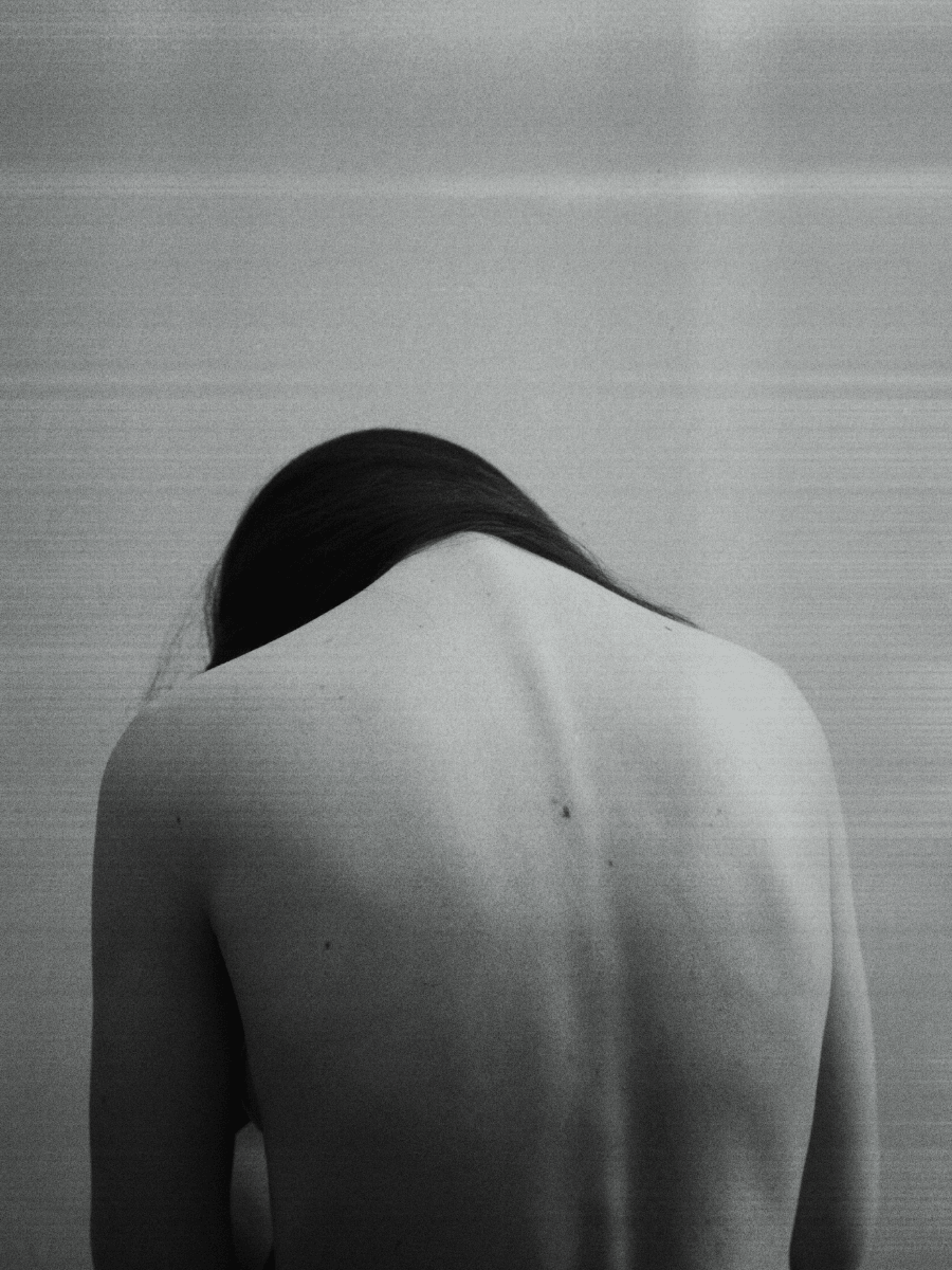 A Libra woman with her bare back and looking down in defeated position against a neutral gray background.