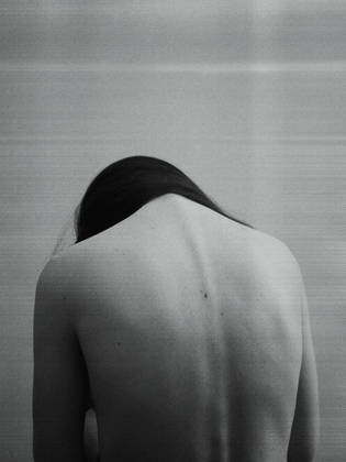 A Libra woman with her bare back and looking down in defeated position against a neutral gray background.