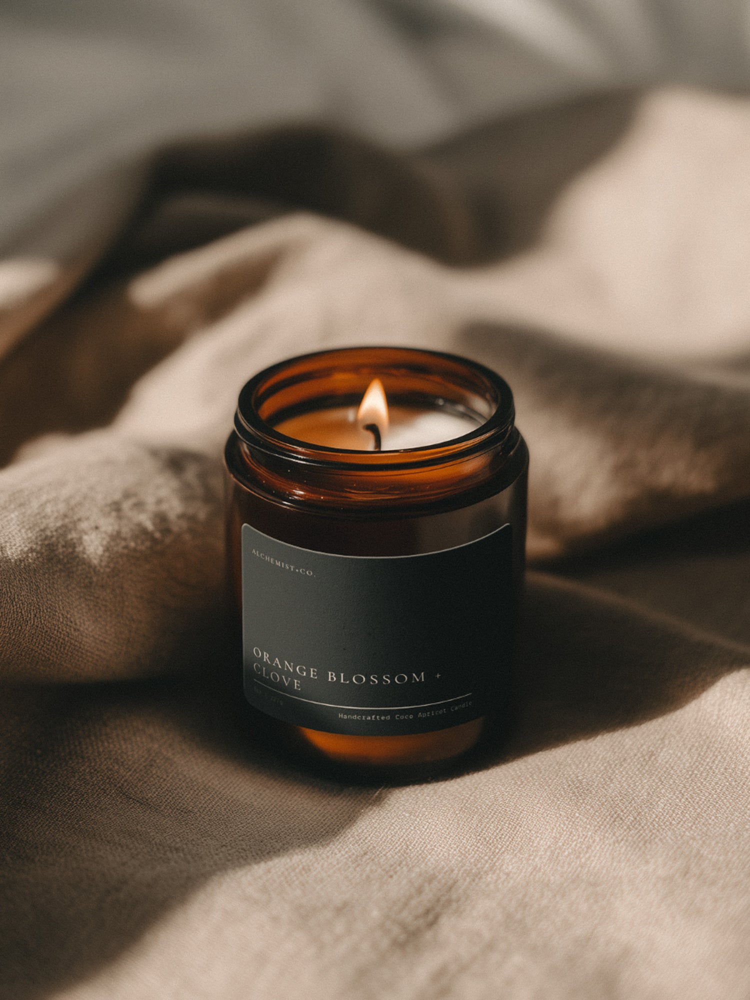 Orange Blossom + Clove Bare candle burning beside soft linen in warm atmospheric light.
