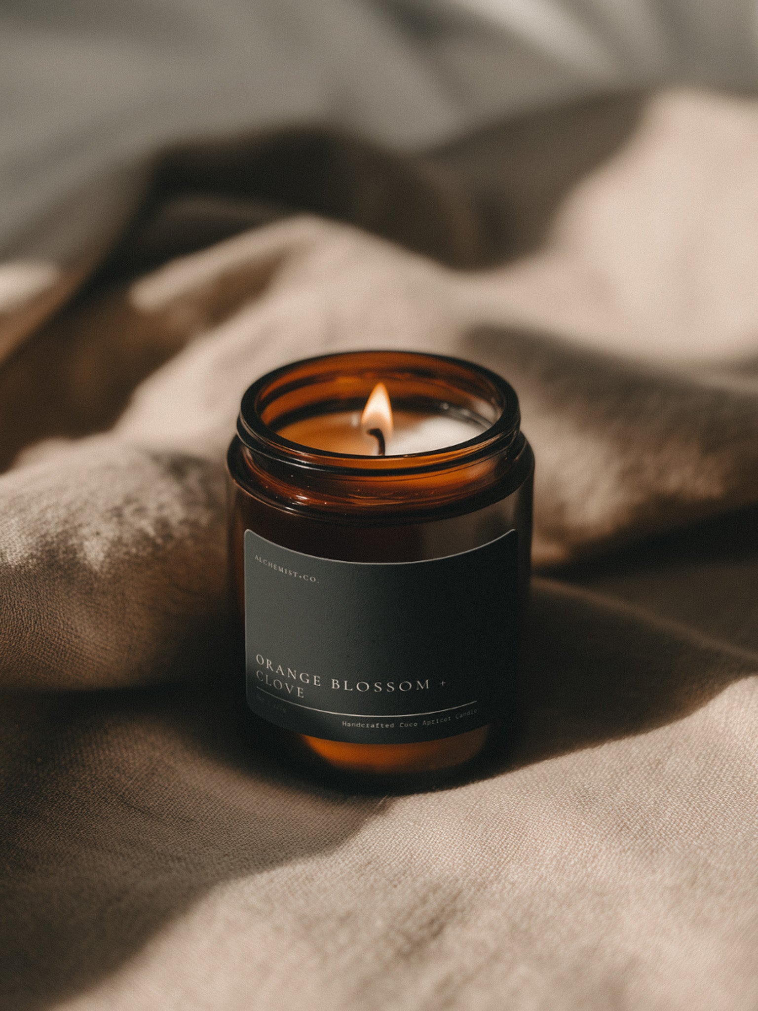 Orange Blossom + Clove Bare candle burning beside soft linen in warm atmospheric light.