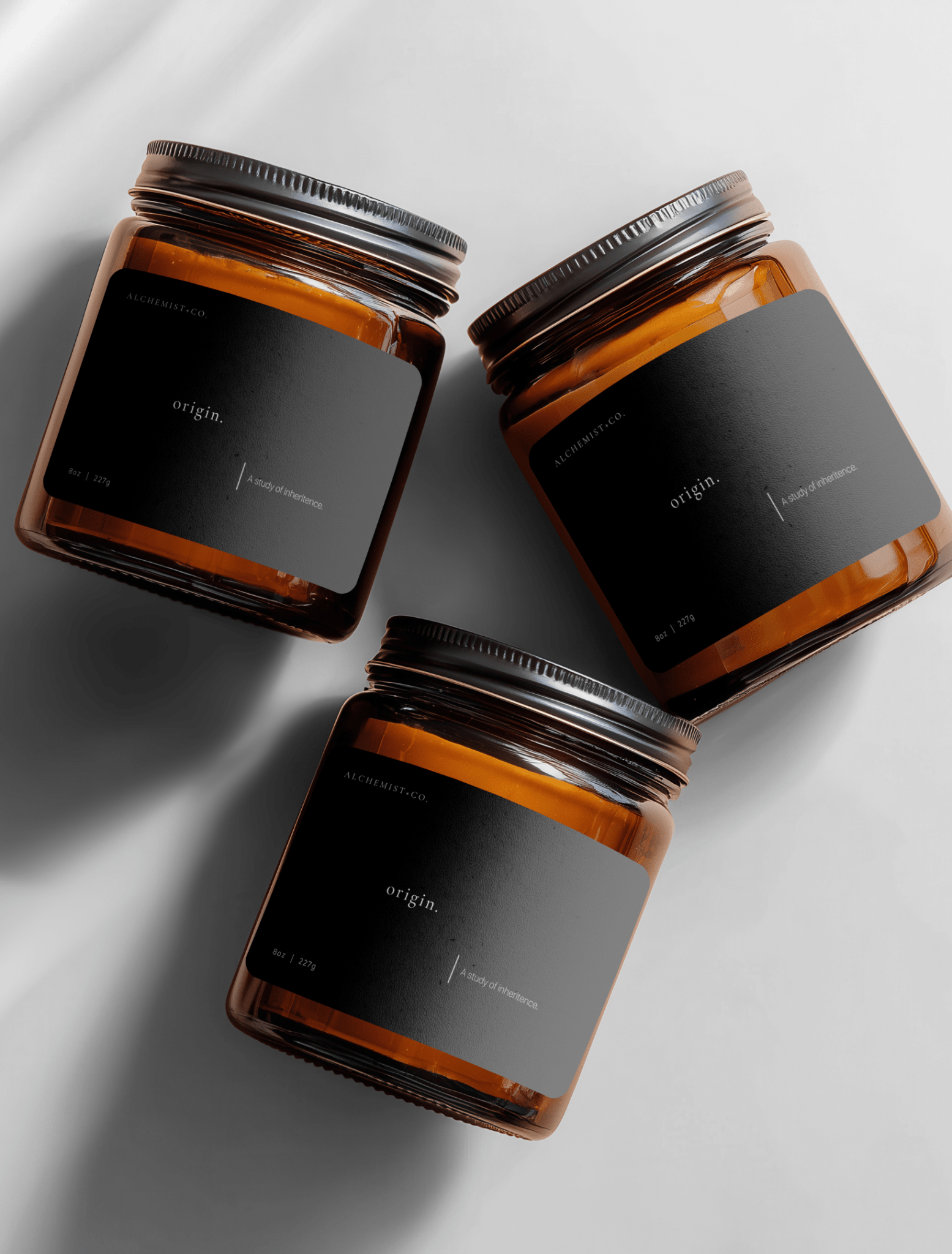 Three amber glass jar candles with black labels and silver lids, arranged diagonally on a light gray background with soft shadows labeled 'Origin'.