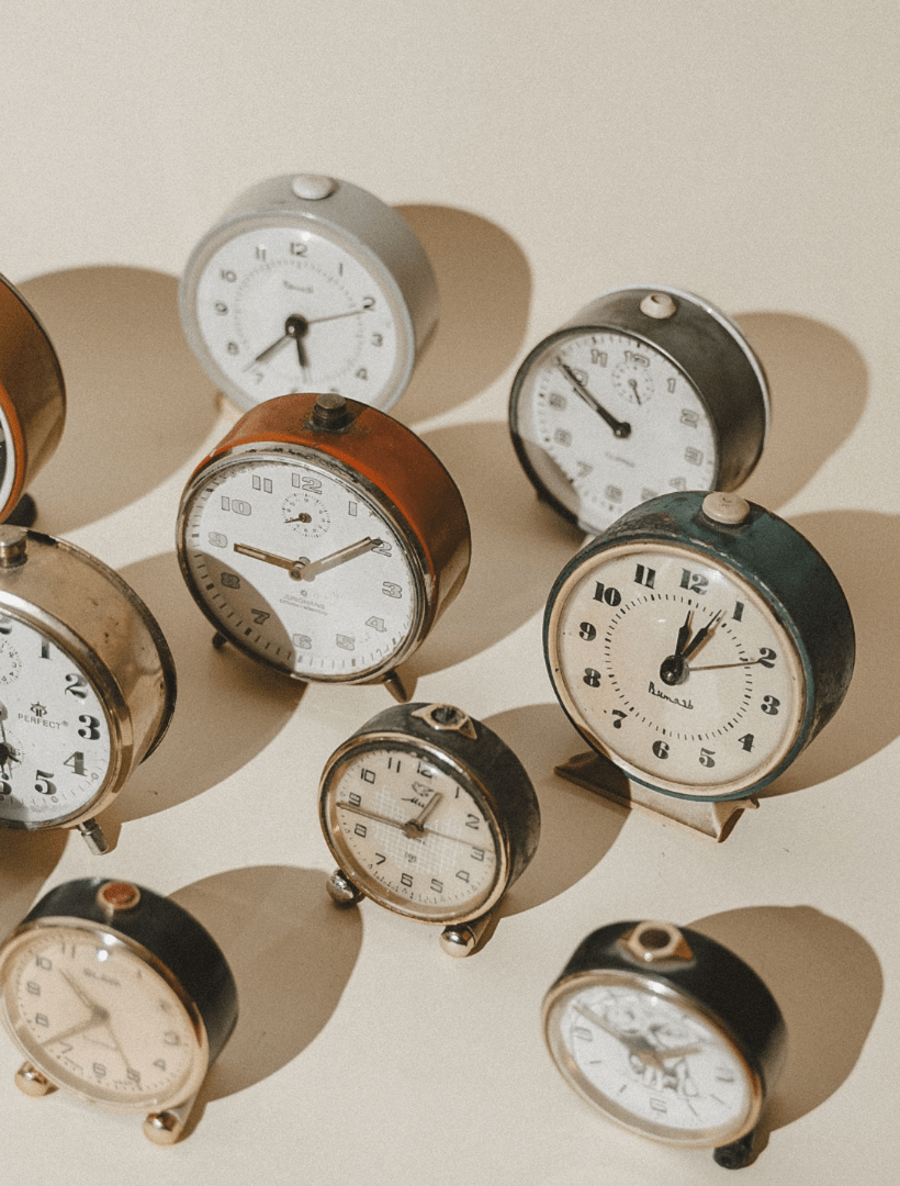 A group of vintage-style tabletop clocks with white faces and varied metal and wood casings, arranged on a warm beige surface with strong directional shadows.