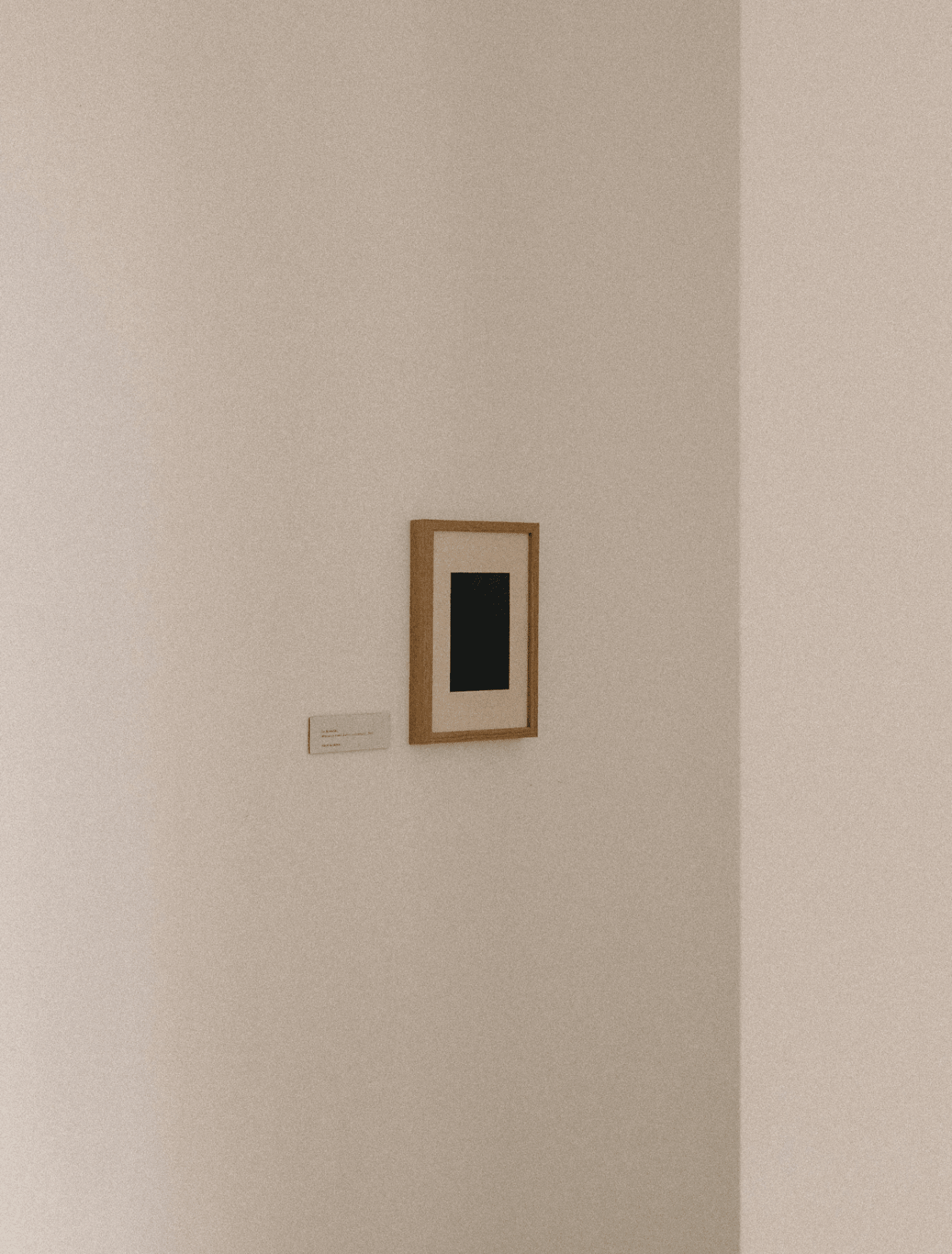 A minimalist framed art print featuring a black square centered within a white mat and a light wood frame, mounted on a textured beige gallery wall with a small label below.