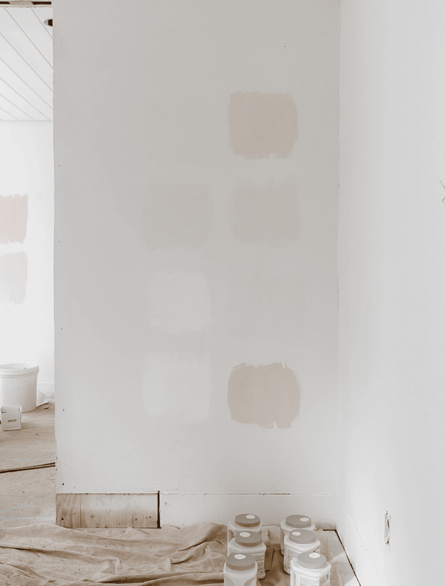A white interior wall with several painted color swatches in soft neutral tones, with paint cans and a drop cloth on the floor nearby.