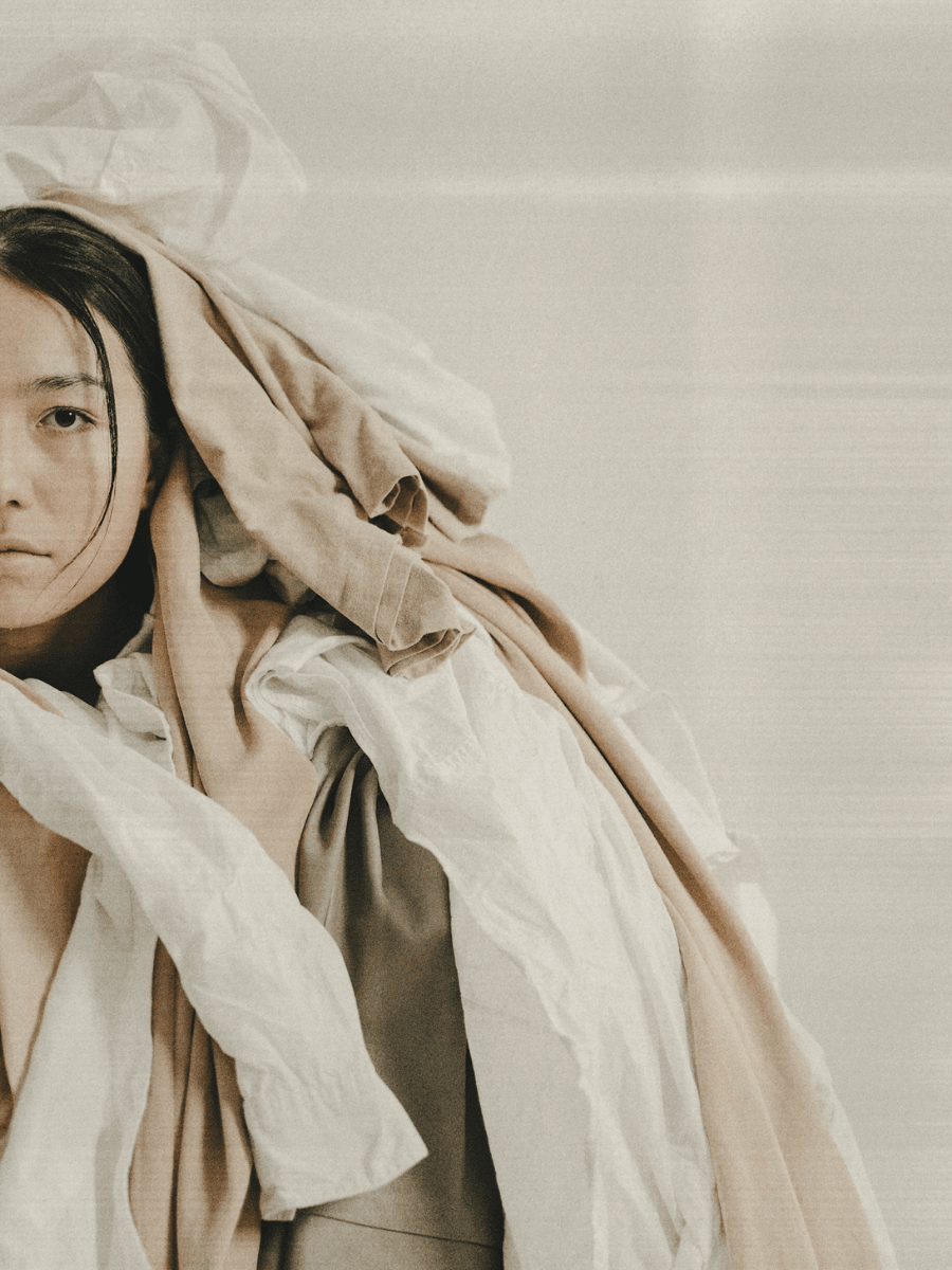 A Gemini woman covered in comfort blankets and clothing on a neutral background.