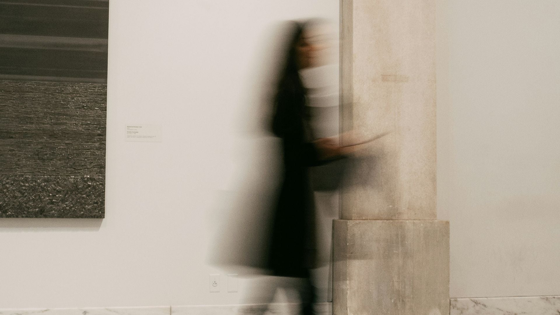 A blurred person in a dark outfit walks past a large concrete column in a minimalist gallery or studio space with white walls and framed artwork.