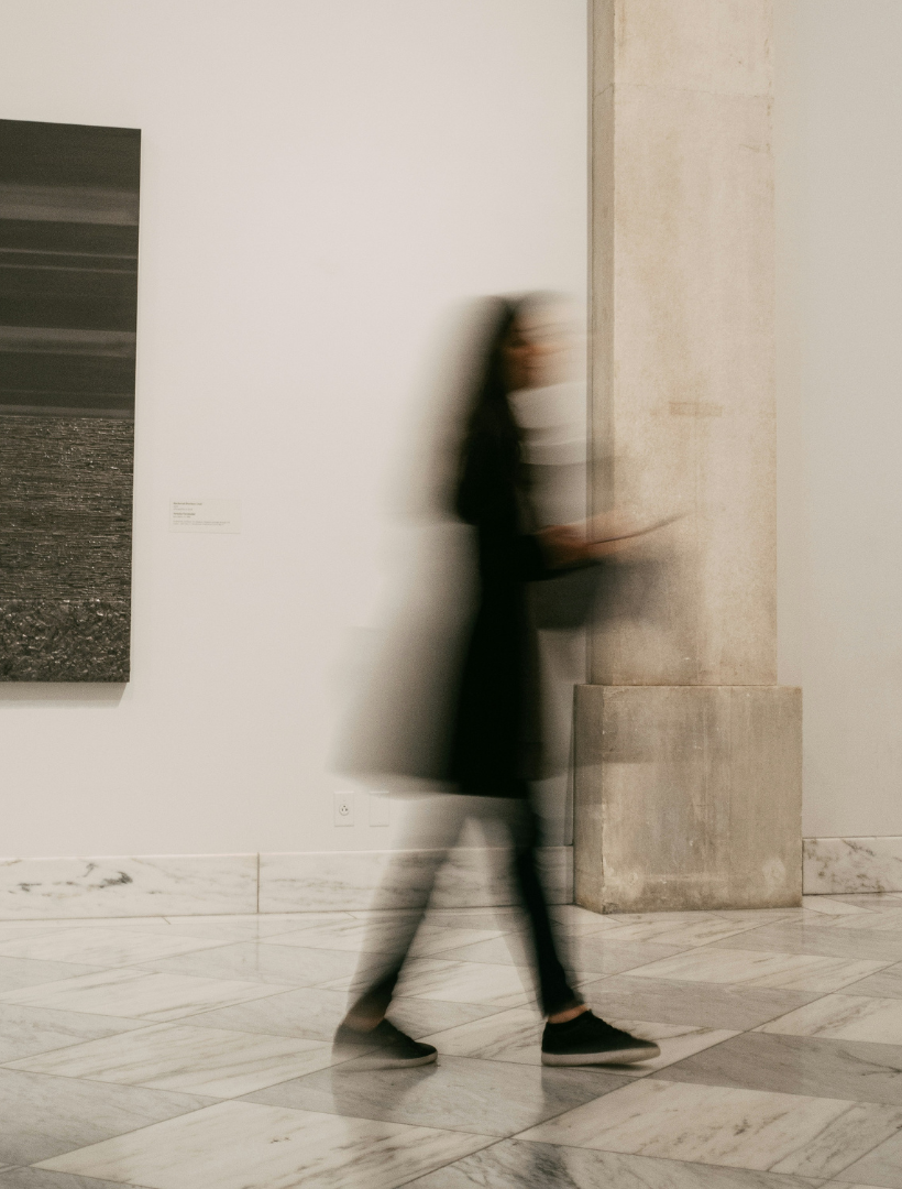 A blurred person walking past a large stone column in a minimalist gallery space with white walls and a marble floor.