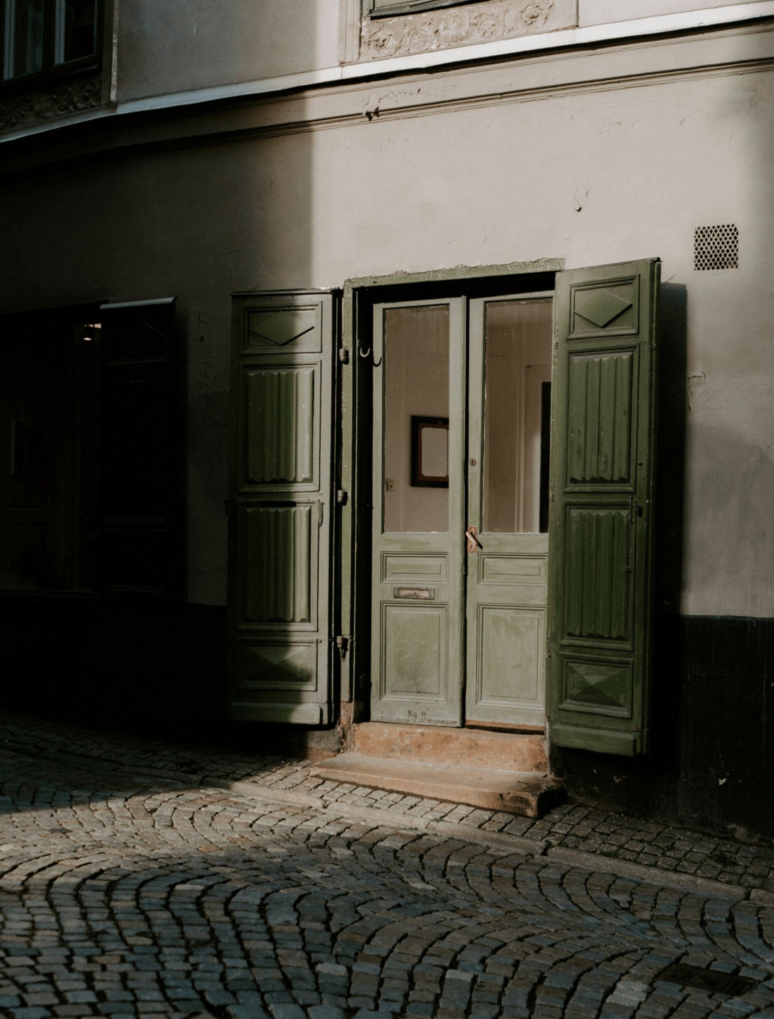 A rustic old building facade with a central double door in a light sage green color, flanked by matching green shutters, set on a cobblestone street under dramatic shadow.