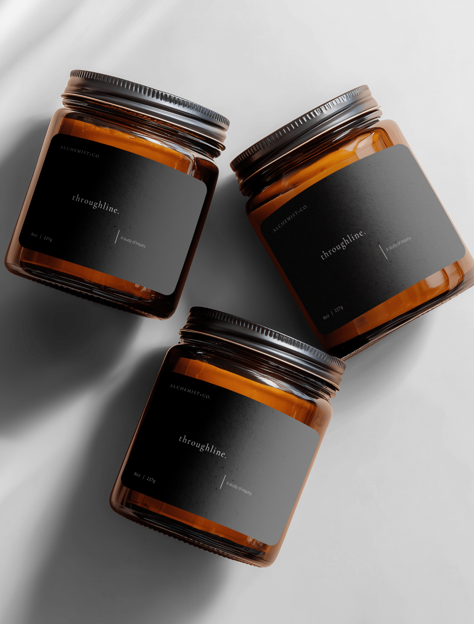 Three amber glass jar candles with black labels and silver lids, arranged diagonally on a light gray background with soft shadows labeled 'Throughline'.