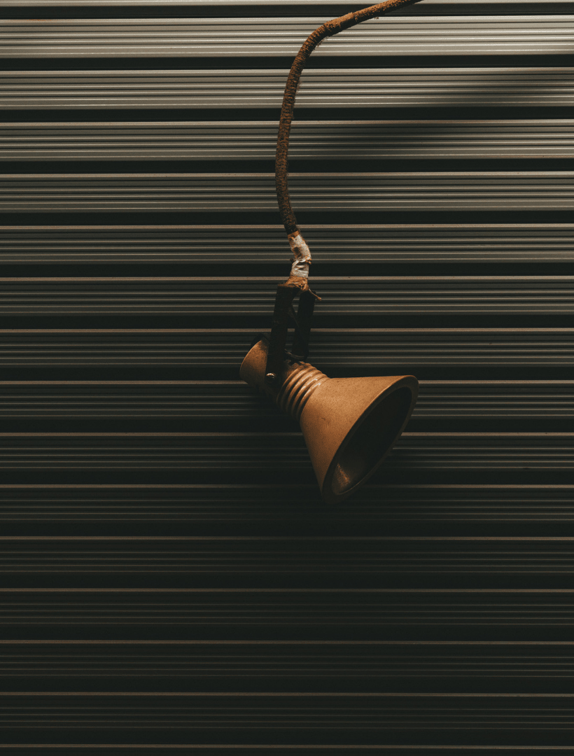A vintage-style metal spotlight with a brown cord, hanging against a dark corrugated metal shutter background.