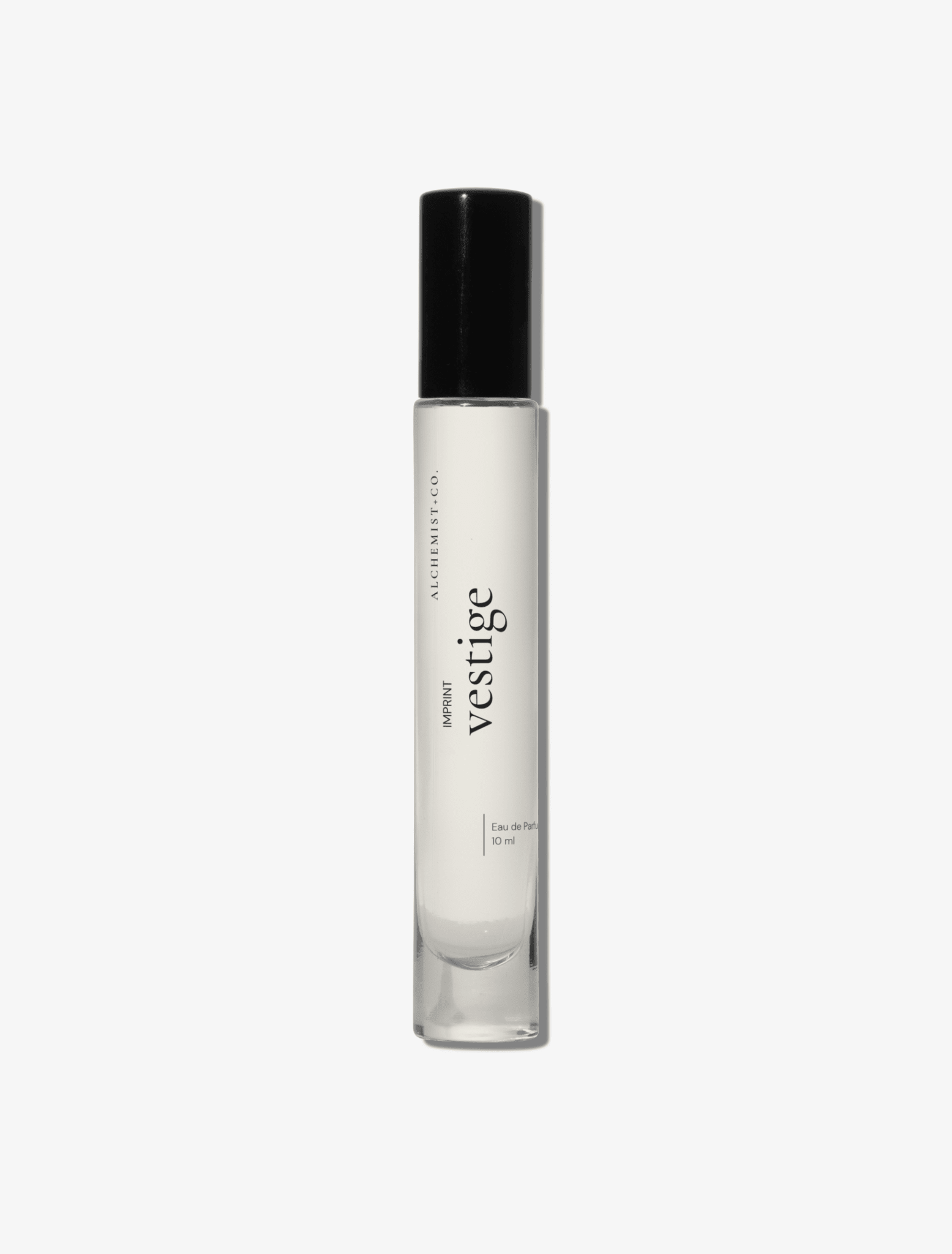 A clear glass perfume bottle with a black cap, featuring a white label with the brand name 'Vestige' by Alchemist + Co.  set against a solid light gray background.
