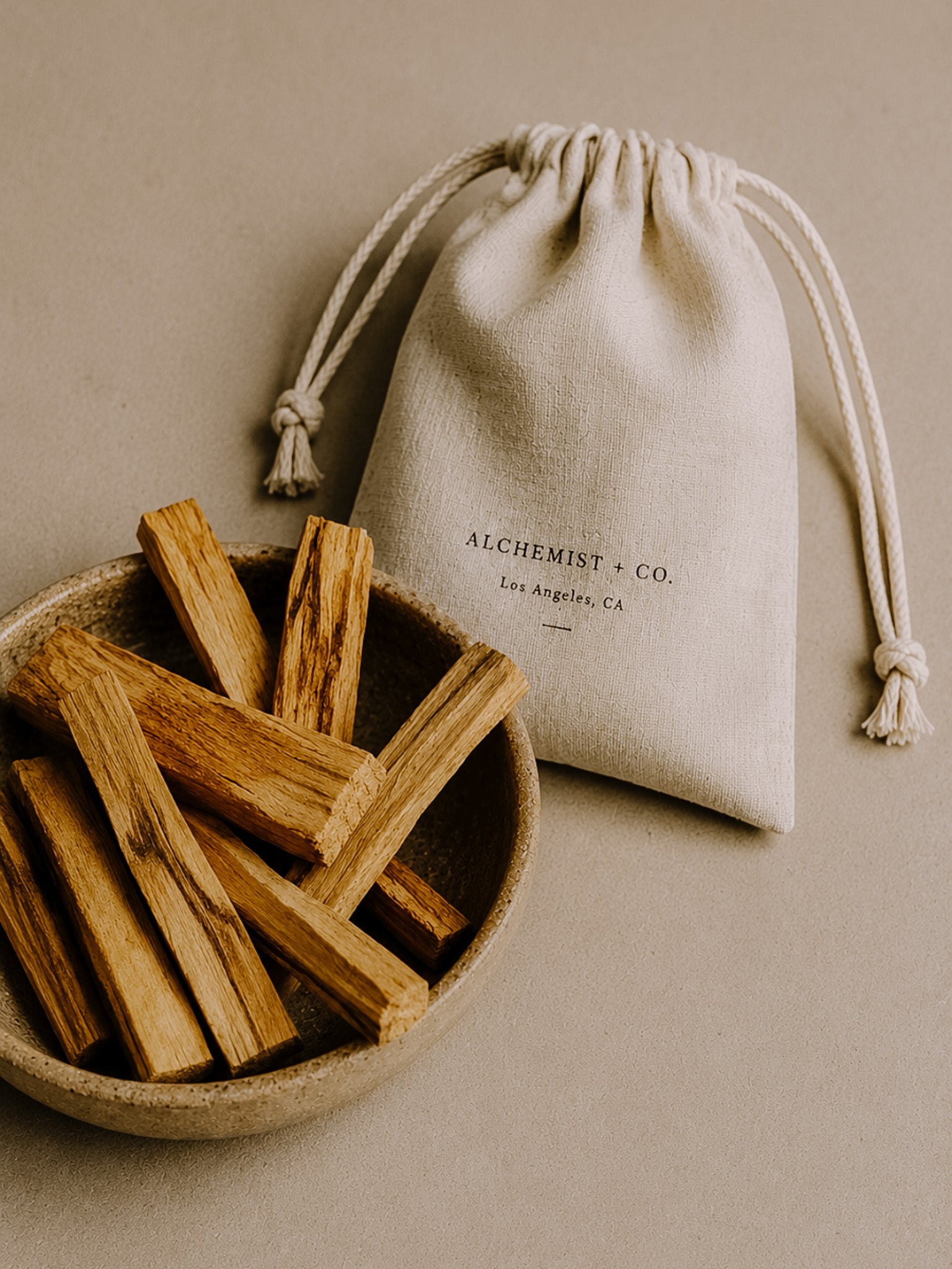 Bowl of Palo Santo sticks with a drawstring bag labeled 'Alchemist & Co.' on a beige background